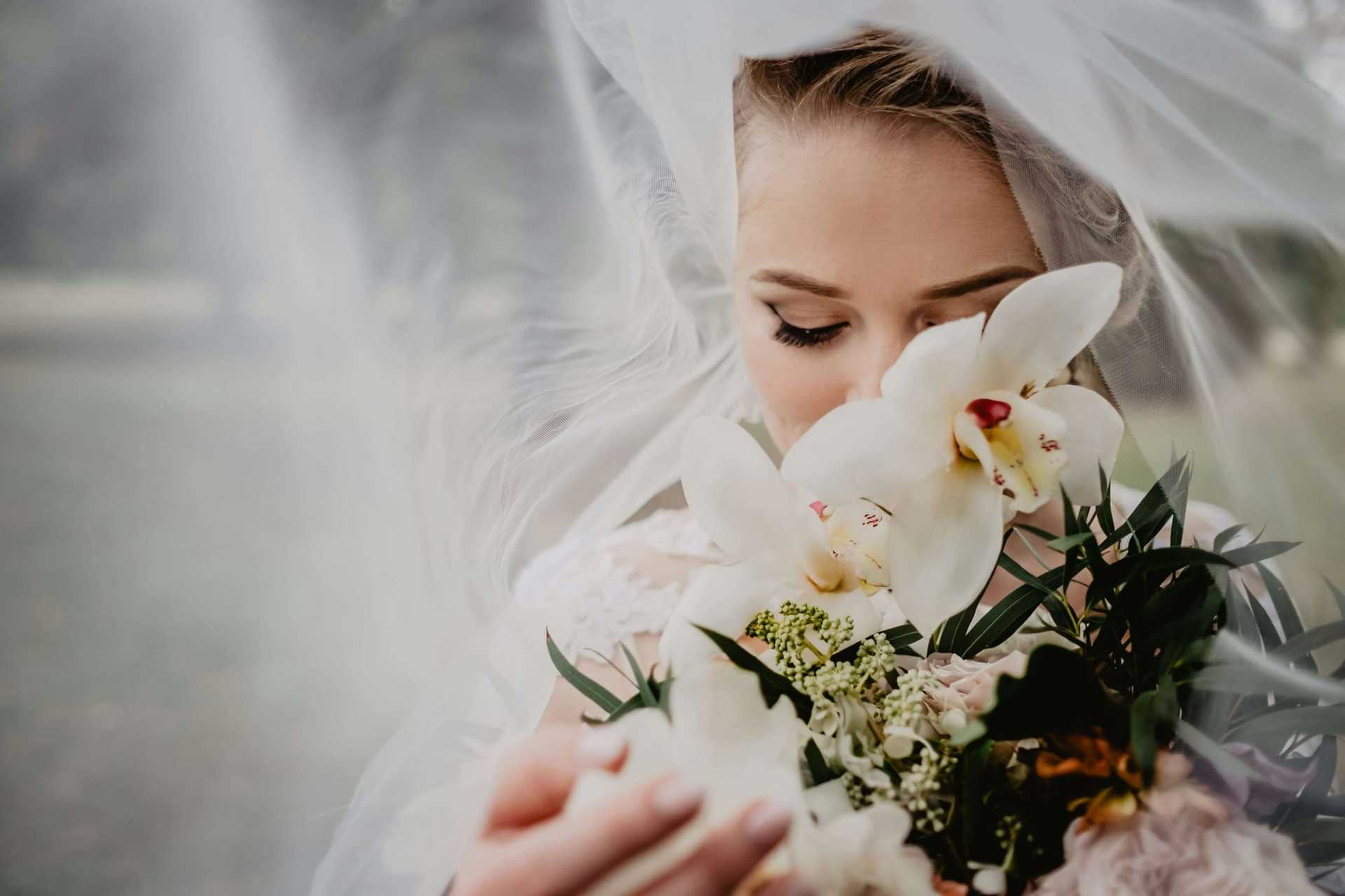 Bride holding white orchids under a veil, eyes closed, in a serene outdoor setting.